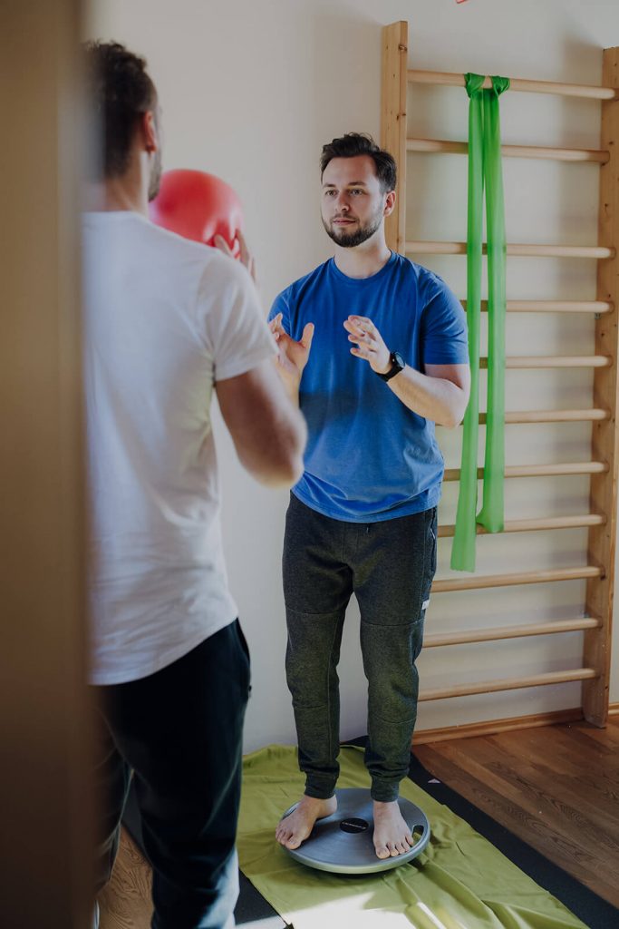 Mann mit blauem Shirt auf Balanceboard blickt auf Mann mit rotem Ball in Händen