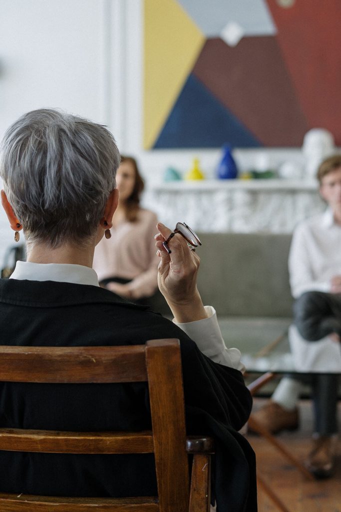 Hinterkopf von Frau mit kurzen grauen Haaren und Ehepaar auf Couch im Hintergrund